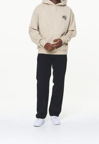 Sudadera beige con bolsillo frontal y logo. Pantalones de pana negros y zapatillas blancas. El modelo está de pie, con las manos al frente.