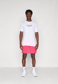 Biely tričko Nike Pro s čiernym logom, ružové šortky, biele tenisky a biele ponožky. Model stojí na neutrálnom pozadí.
