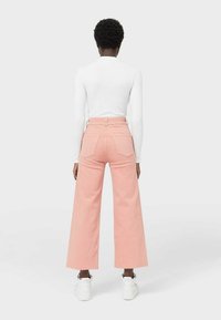 Pantaloni a gamba larga rosa chiaro con vita alta, dotati di tasche posteriori e chiusura con zip. Abbinati a un top aderente a manica lunga bianco.