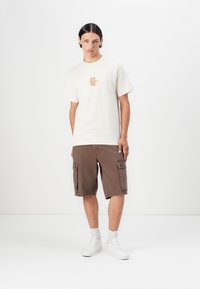 Kortermet kremfarget T-skjorte med gul grafisk design, kombinert med brune cargo-shorts med sidelommer. Hvite joggesko fullfører antrekket.
