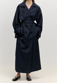 Mörkblå trenchcoat med vita ränder, dubbelsidig design, breda revers och långa ärmar. Inkluderar ett matchande bälte och fickor.