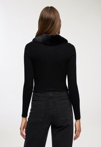 Kobieta z długimi brązowymi włosami nosi czarny sweter z prążkowanego materiału z kołnierzem z sztucznego futra oraz czarne dżinsy z wysokim stanem, pokazana z tyłu na białym tle.