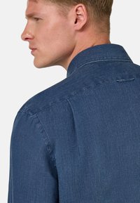 Temno moder jeans srajca s ovratnikom, vidnim šivom, strukturirano tkanino in enim zadnjim plišem za olajšanje gibanja.