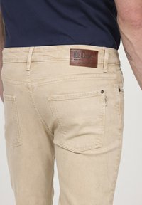 Jeans en denim beige avec une coupe slim, dotés de poches arrière, d'une étiquette en cuir et de coutures contrastées. Texture lisse et détails de design minimal.