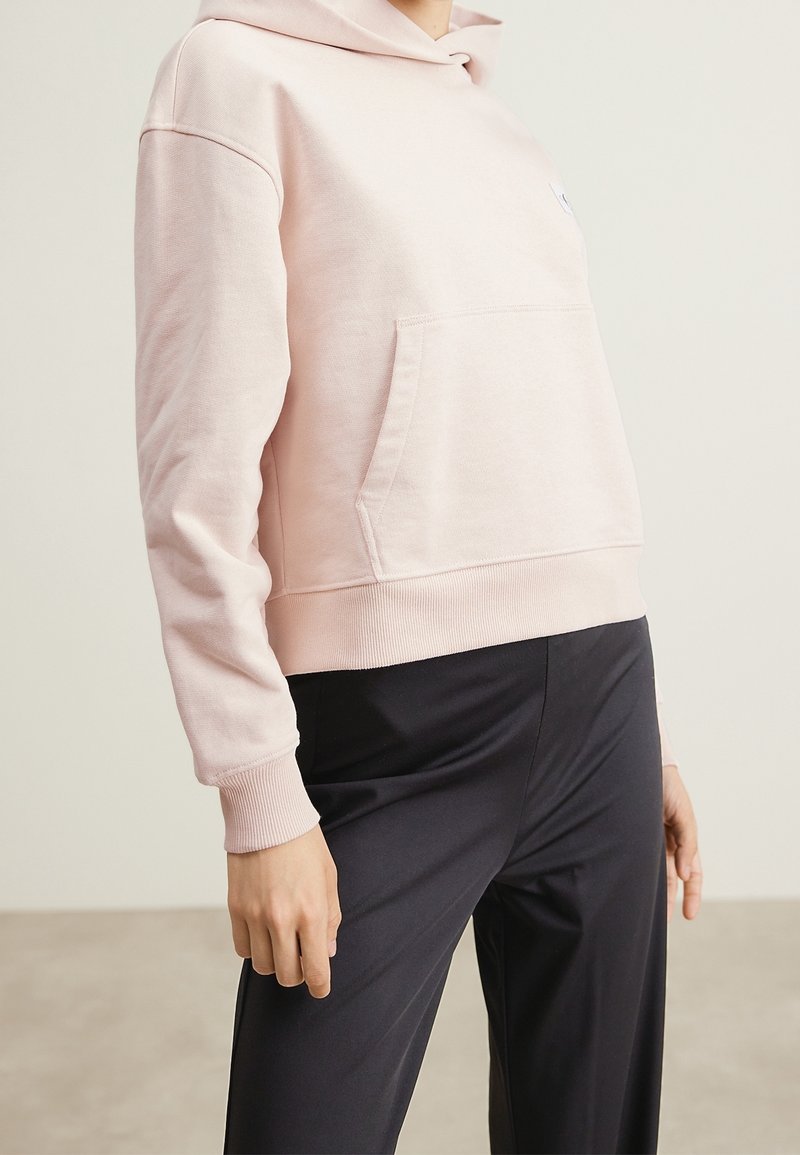 Personne portant un sweat à capuche rose clair avec une poche avant et un pantalon noir, debout à l'intérieur devant un fond uni.