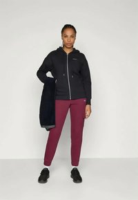 Schwarze Zip-Up Kapuzenjacke mit verstellbarem Kordelzug, kombiniert mit burgunderfarbenen Jogginghosen. Das Outfit umfasst ein dunkles Handtuch und schwarze Turnschuhe.