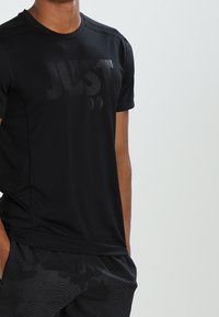 Camiseta deportiva negra con tela texturizada y transpirable. Presenta un gráfico sutil de "JUST DO IT" en un tono más oscuro. Manga corta y cuello redondo.