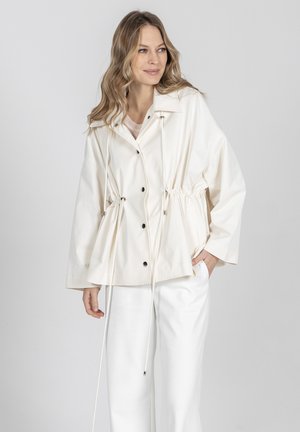 Kobieta ubrana w luźną kremową kurtkę z troczkami i białe spodnie, pozująca na tle jednolitego jasnoszarego tła.