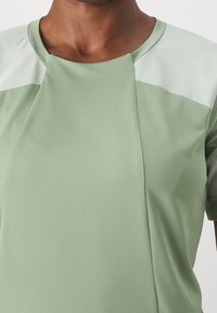 Vaaleanvihreä urheilupaita, jossa on rakenteellinen kangas ja sileät, vaaleammassa sävyssä olevat vastaväriset hartiapaneelit. Pyöreä kaula-aukko.