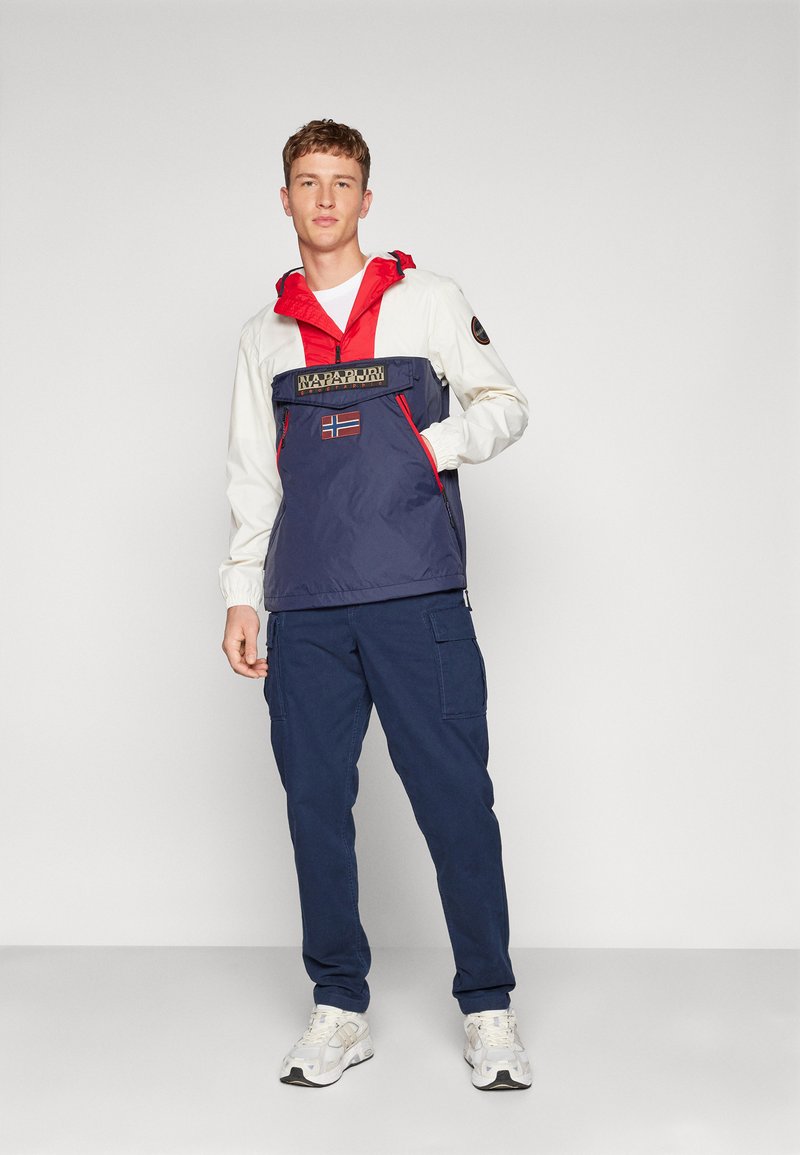 Giovane uomo che indossa una giacca Napapijri bianca, rossa e blu navy con un patch della bandiera norvegese, pantaloni cargo blu navy e sneaker bianche, in piedi davanti a uno sfondo neutro.