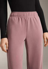 Világos rózsaszín melegítőnadrág elasztikus derékrésszel, sima textúrával és laza szabással, fekete hosszú ujjú felsővel párosítva.