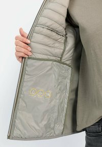 Intérieur d'une veste rembourrée gris clair avec une texture lisse. Présente des icônes jaunes indiquant qu'elle est déperlant, coupe-vent et sans duvet.