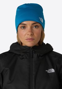 Jasnoniebieska czapka beanie z gładkiego materiału, z białym logo The North Face, noszona z czarną kurtką zapinaną na zamek z wysokim kołnierzem.