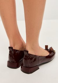 Ballerines en cuir vernis bordeaux avec un talon géométrique bas, ornées d'un nœud décoratif avec des accents imprimé léopard. Texture lisse.