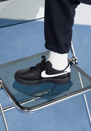 Person iført sorte Nike-sneakers med hvide sokker og sorte bukser, der hviler foden på en glas- og kromstol på en blå gulv.