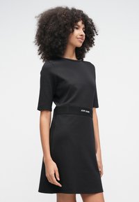 Frau mit lockigem Haar, die ein schwarzes kurzes Kleid mit einem markierten Taillenband trägt und vor einem schlichten hellen Hintergrund steht.