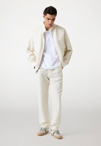 Beige bomber jacket with a zip, white t-shirt underneath, and matching beige pants. Gray and green sneakers with a gum sole complete the outfit.