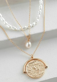 Collier à trois couches composé d'une chaîne dorée, d'un pendentif rond texturé et d'une corde ornée de perles, comprenant des perles de formes irrégulières.