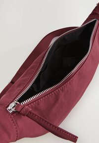 Burgunderfarvet crossbody taske med en lynlåsåbning, der har en glat overflade, sort stofforing og en flad rem for nem bæring.