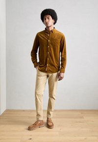 Bruine corduroy button-up shirt met borstzak, beige slim-fit broek en bruine loafers, geplaatst op een houten vloer met een neutrale achtergrond.