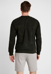 Uomo che indossa una felpa scura con motivo camouflage e righe decorate sulle maniche, pantaloni corti grigi chiari, in piedi di spalle su uno sfondo bianco.