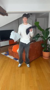 Jonge man lacht en houdt een grijs shirt en een zwarte trui vast in een woonkamer met een houten vloer en een oranje bank.