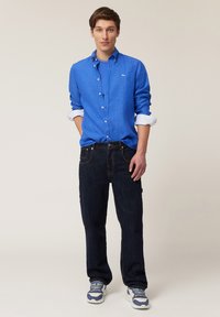 Camisa de lino azul llevada sobre una camiseta azul, combinada con jeans de mezclilla oscuros y zapatillas blancas con acentos azules.