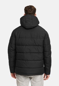 Mann in schwarzer Daunenjacke mit Kapuze, mit dem Rücken zur Kamera, kombiniert mit hellgrauen Jeans, vor einem schlichten Hintergrund stehend.