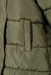 Olīvu zaļš piešūta jaka ar kabatu, horizontālas šuves un flaps, kas sedz atveri.