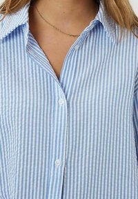 Chemise à rayures bleu clair et blanches avec col, fabriquée en tissu doux, présentant une coupe décontractée et des boutons visibles.