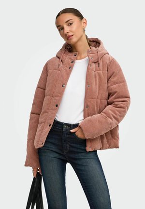 Femme portant une doudoune en velours côtelé rose par-dessus une chemise blanche et un jean bleu, tenant un sac à main noir sur un fond uni.