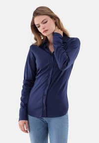 Giovane donna in camicia azzurra a maniche lunghe e jeans chiari, con una mano che tocca il collo, che guarda in basso.