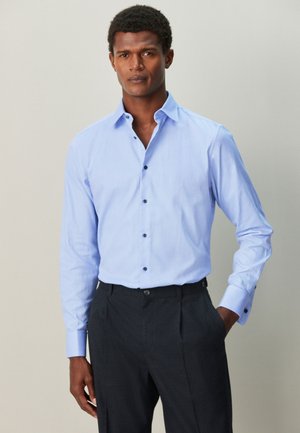Hombre que lleva una camisa azul clara con botones oscuros y pantalones oscuros, de pie sobre un fondo liso.
