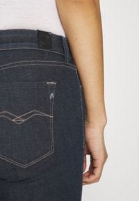 Vaqueros de mezclilla azul oscuro con costuras visibles, un bolsillo trasero con un patrón de ola y una etiqueta de cuero negro en la cinturilla.