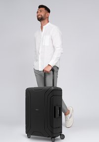 Man in wit shirt en grijze broek staan met één been omhoog, terwijl hij een zwarte rolkoffer aan de handgreep vasthoudt tegen een effen achtergrond.