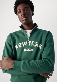 Sweatshirt vert zippé au quart avec « NEW YORK » et « SCHOTT » imprimés en crème. Le matériau semble doux avec des poignets et un col côtelés.