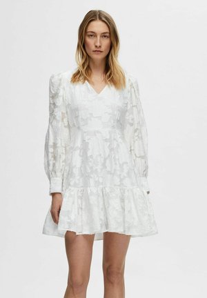 Robe blanche avec une texture florale transparente, manches longues bouffantes, décolleté en V et ourlet en couches, créant une silhouette fluide.