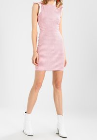 Robe ajustée sans manches à motifs carreaux rouges et blancs, avec des détails de volants aux épaules. Associée à des bottines blanches. 