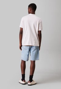 Polo shirt a maniche corte grigio chiaro, vestibilità rilassata, abbinato a pantaloni corti in denim blu chiaro. Le scarpe sono di colore crema con accenti scuri e calze nere.
