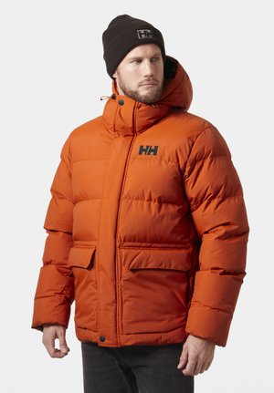 Man draagt een oranje gewatteerd jack met capuchon en een zwarte muts, kijkt opzij tegen een effen achtergrond.