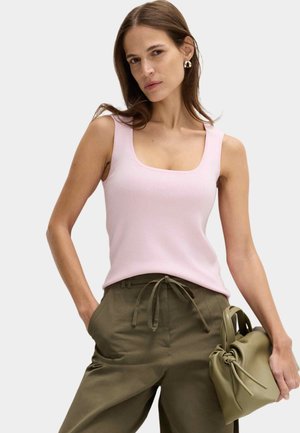 Femme portant un haut sans manches rose clair et un pantalon vert olive, tenant un sac à main assorti vert olive dans la main droite.