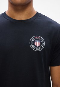 Must musta T-särk, millel on ümar logo rinnal ja tekst "GANT 1949 NEW HAVEN CONNECTICUT" ning punased, valged ja sinised aktsendid.
