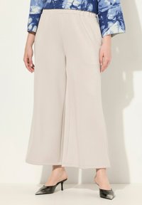 Pantalons larges beige clair en tissu doux avec une taille élastique, associés à des mules noires à talons, mettant en valeur le fluide des pantalons.