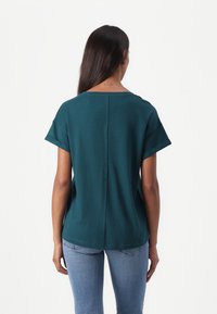 Teal kortermet topp med rund halslinning og en midtseamsdetalj bak. Kombinert med lyseblå jeans, fremstår stoffet som mykt.