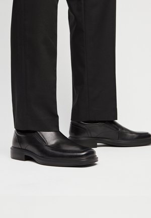 Scarpe eleganti in pelle nera senza lacci indossate con pantaloni formali neri, mostrate dalla caviglia in giù su uno sfondo chiaro.
