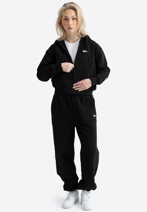 Mujer con sudadera negra con cremallera y logo de león, pantalones deportivos negros a juego, camiseta blanca y zapatillas blancas, de pie frente a fondo blanco.