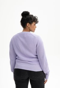 Lavendelfarbenes Strickpullover mit strukturiertem Muster, langen Ärmeln und rundem Ausschnitt, kombiniert mit schwarzen Jeans. Sichtbares geripptes Saumdetail.