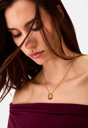 Collana d'oro con un pendente geometrico, indossata sulla pelle nuda, abbinata a un top a spalle scoperte di un profondo bordeaux. Texture liscia, design minimalista.