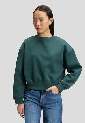 Donna con capelli lisci e scuri che indossa una felpa verde scuro e jeans blu, in piedi con una mano in tasca davanti a uno sfondo semplice.