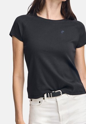 Kvinde iført en sort kortærmet trøje, let stoppet ind i hvide jeans med sort bælte foran en ensfarvet baggrund.